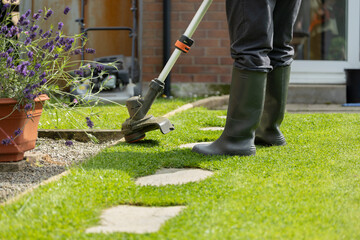 Woman in black trouser pants and wellie boots using electric strimmer weed whacker to trim grass...