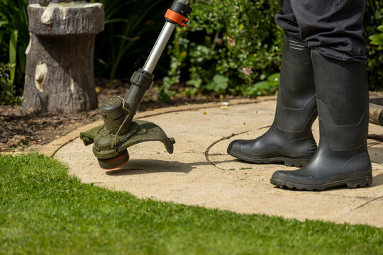 Woman in black trouser pants and wellie boots using electric strimmer weed whacker to trim grass lawn edge