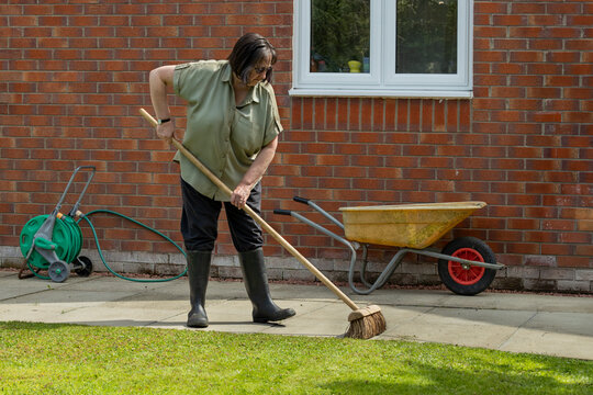 Woman in black trouser pants and wellie boots sweeping concrete slab paved patio after trimming garden lawn