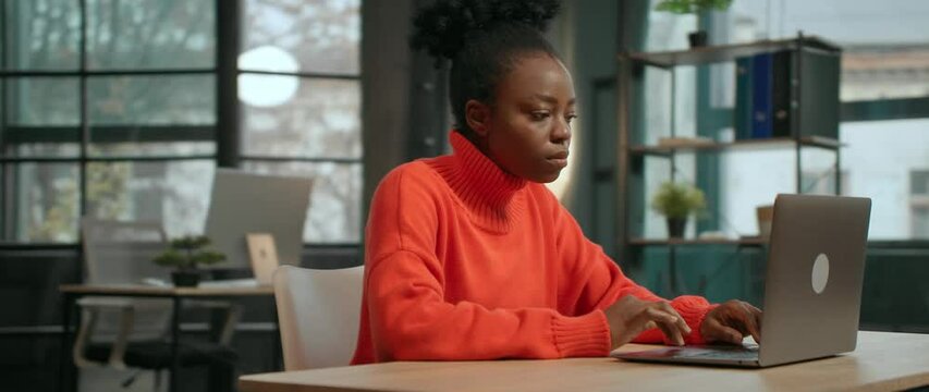 Parralax effect. 360 degree shot of good-looking friendly African American woman using silver laptop while sitting at desk. Focused girl in orange sweater browsing on Internet. Tracking moving around