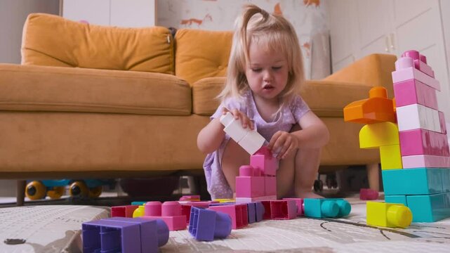 A cute child toddler girl in a dress is sitting on the carpet in the room and playing with a colorful constructor.