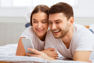 Fototapeta premium Mobile App. Cheerful Couple Using Smartphone Lying In Bed At Home In The Morning. Selective Focus