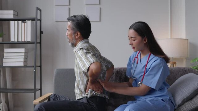 Nurse wearing blue scrubs examines a senior man experiencing back pain while sitting on a couch