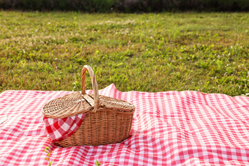 Picnic wicker basket with napkin and red checkered blanket on green grass outdoors, space for text