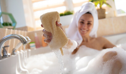 A woman in a towel enjoys a relaxing bath, playfully squeezing a sponge towards the camera, with water dripping down. The bright bathroom and soft bubbles create a refreshing, carefree atmosphere.