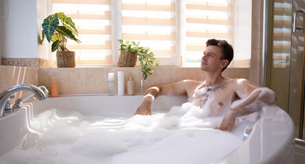 Handsome man enjoys a relaxing bubble bath in a sunlit bathroom. He leans back contentedly, surrounded by plants and bath products, conveying a sense of calm and self-care in a modern, cozy setting