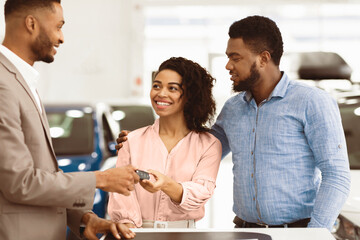 Car Dealer Giving Key To Cheerful Afro Couple Buyers Standing In Auto Dealership Center. Cars Sales. Selective Focus