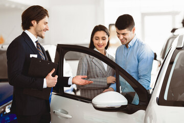 Auto Business Concept. Handsome Assistant Presenting New Luxury Auto With Open Door To Young Couple
