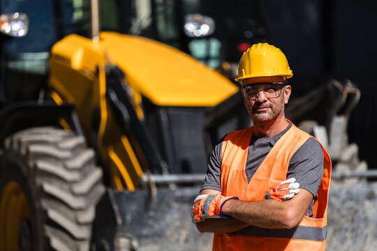 Construction worker in safety gear stands confidently near a yellow heavy machinery during daylight hours at a job site