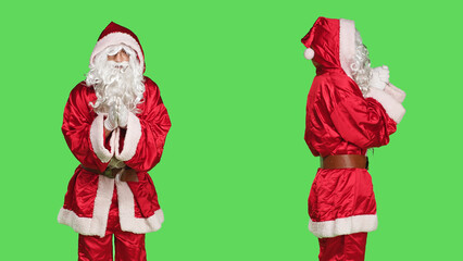 Faithful santa claus praying in studio, holding hands in a prayer and standing against greenscreen backdrop. Model portraying father christmas with festive red costume being religious.