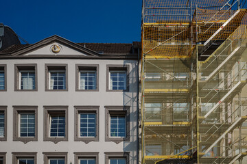 A building undergoing renovation. On the left, a well-preserved section of the structure shows classic architectural elements, while on the right, scaffolding is set up for repair work.