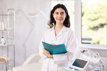 Female cosmetic doctor meticulously stands in her clinic, recording detailed notes in folder,...