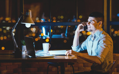 Business Call. Cheerful Businessman Talking On Cellphone Communicating With Distant Colleague Sitting At Workplace Indoor At Night