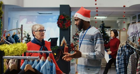 Employee assisting older woman with picking right shirt to give as present to husband for upcoming Christmas holiday season. Client browsing through clothes in xmas adorn fashion shop