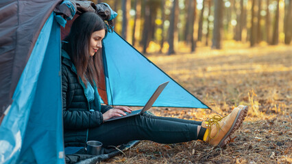 Stay connected. Young girl using laptop in camping tent, hiking in autumn forest, empty space