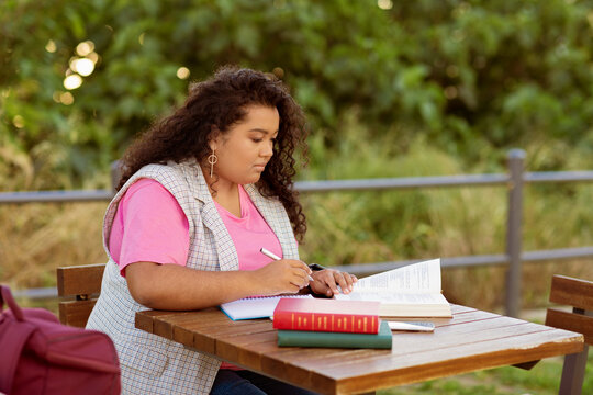 A student engaged in studying while seated at a wooden table outdoors. The individual writes in a notebook, with books beside them, immersed in their studies on a sunny day.