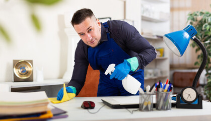 Cleaning man in overalls wipes dust from table in the office