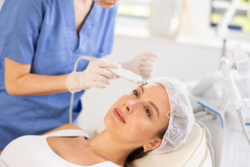 Young woman cosmetologist performs hardware facial microcurrent procedure to young female patient