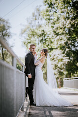 Naklejka premium A bride in a lace gown lovingly touches the groom’s face on a sunny day while standing together on a bridge.