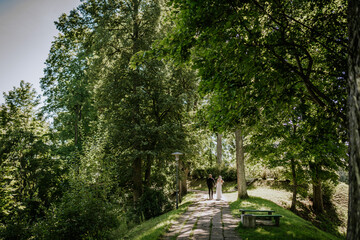 Obraz premium A bride and groom hold hands in a peaceful, sunlit forest path surrounded by tall trees and lush greenery during their wedding day.