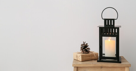 Black lantern with burning candle, gift and fir cone on stool near light wall