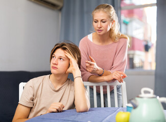 Frowning teenager with bored expression listening to reprimanding from his mother at home..