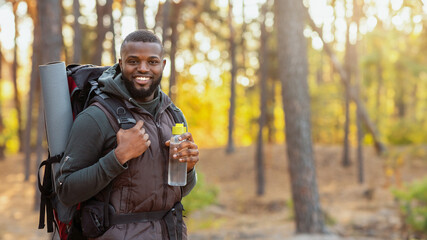 Smiling young african man wearing backpack standing over autumn forest background, holding bottle of water, copy space