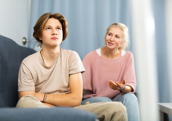 Upset teenager boy after quarrel with his mother in domestic interior, mother consoles son