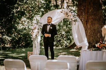 A groom in a black tuxedo stands under a floral wedding arch in an outdoor ceremony with greenery...