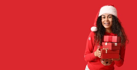 Young African-American woman in Christmas sweater and Santa hat and with gifts on red background with space for text