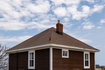 A vintage four sided brown wooden house with an asphalt shingled hip roof. There's a red brick...