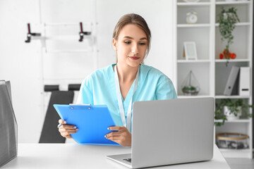 Female medical intern with clipboard using laptop at hospital