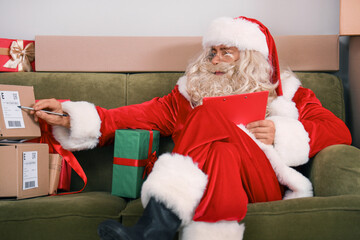 Naklejka premium Santa Claus with clipboard checking list of parcels while sitting on sofa in living room