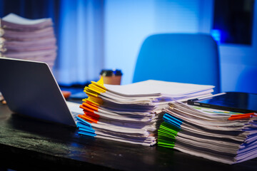 stack of papers rests on a black wooden desk in a dimly lit office room. The blue lighting adds a...