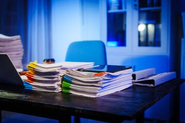 stack of papers rests on a black wooden desk in a dimly lit office room. The blue lighting adds a...