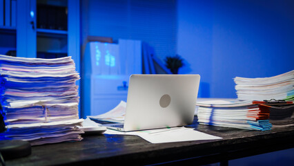stack of papers rests on a black wooden desk in a dimly lit office room. The blue lighting adds a...