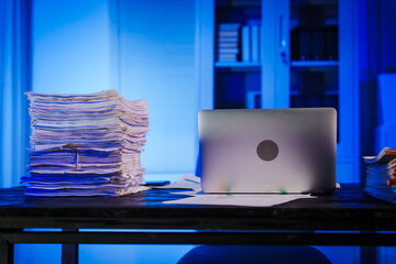 stack of papers rests on a black wooden desk in a dimly lit office room. The blue lighting adds a...