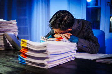 A businessman works late into the night, facing a stack of papers on a black wooden desk. The blue lighting reflects the intensity of his overtime efforts in a busy office environment.