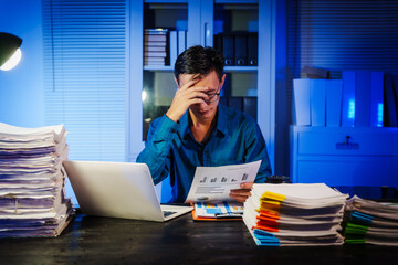 Asian businessman works late into the night, focused on a stack of papers on his black wooden desk. The office is dimly lit with a blue glow, reflecting the busy atmosphere of overtime.