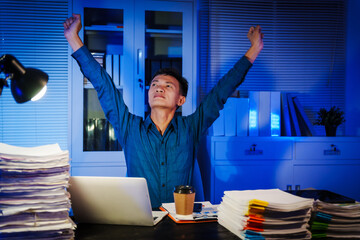Asian businessman works late into the night, focused on a stack of papers on his black wooden desk....