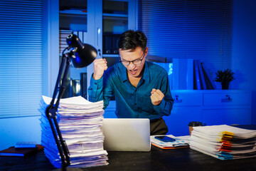 Asian businessman works late into the night, focused on a stack of papers on his black wooden desk....