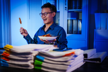 Asian businessman works late into the night, focused on a stack of papers on his black wooden desk. The office is dimly lit with a blue glow, reflecting the busy atmosphere of overtime.