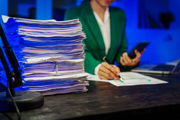 Asian businesswoman works late into the night in a busy office, facing a stack of papers on a black wooden desk, with a laptop and mobile nearby under soft blue lighting.