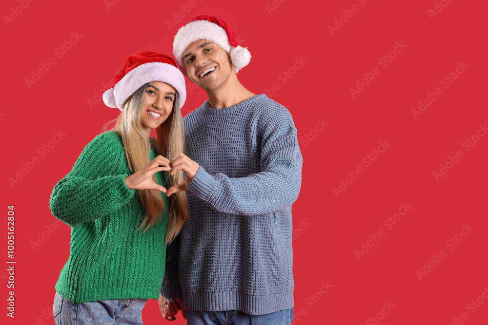 Sticker Happy young couple in Santa hats making heart gesture on red background