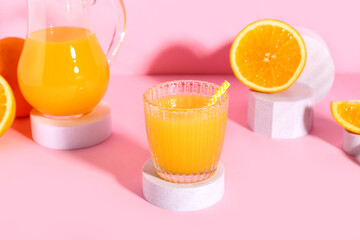 Decorative podiums with glass and jug of fresh orange juice on pink background