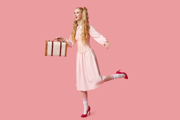Beautiful young happy woman dressed as doll with suitcase on pink background