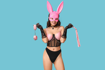 Beautiful young Asian woman in sexy lingerie with bunny mask holding whip and handcuffs on blue...
