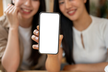 A close-up of a woman showing her smartphone to the camera while sitting indoors next to her friend.