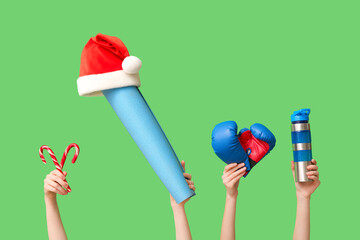 Female hands holding sport bottle, boxing gloves, candy cane, yoga mat and Santa hat on green background. Christmas sport composition