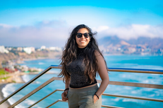 Young Latina woman smiling and enjoying the ocean view on a sunny day in Larcomar, Miraflores, Lima, Peru, with the coastline and mountains in the background.